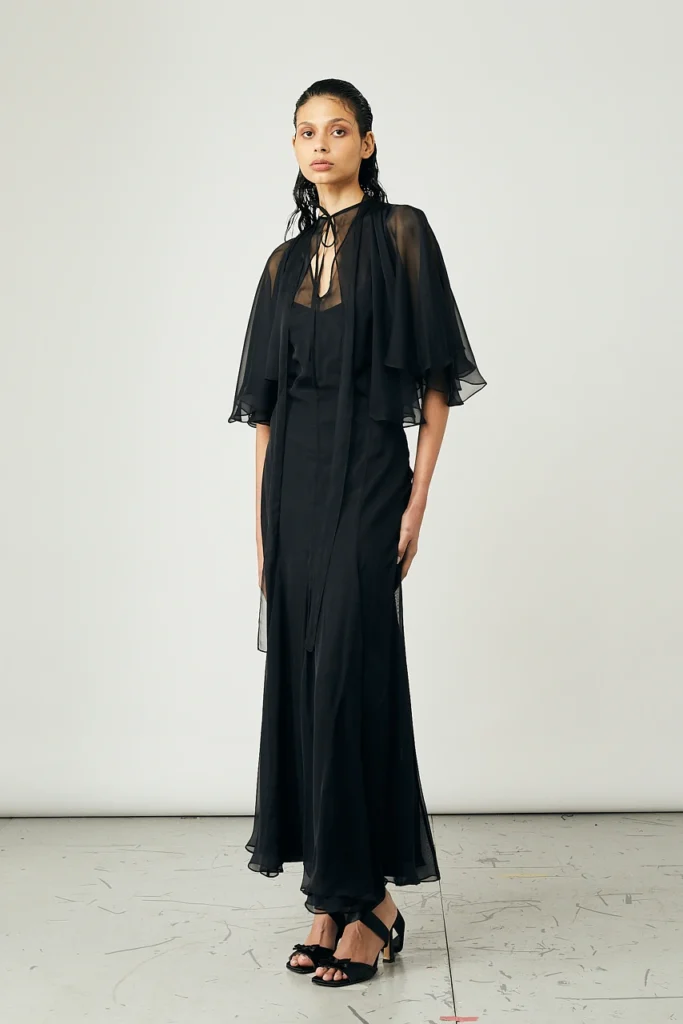Portfolio Lorenzo Berni_094 - Lorenzo Berni Model in a full-length black gown with a sheer cape and tie at the neck, standing against a neutral backdrop.