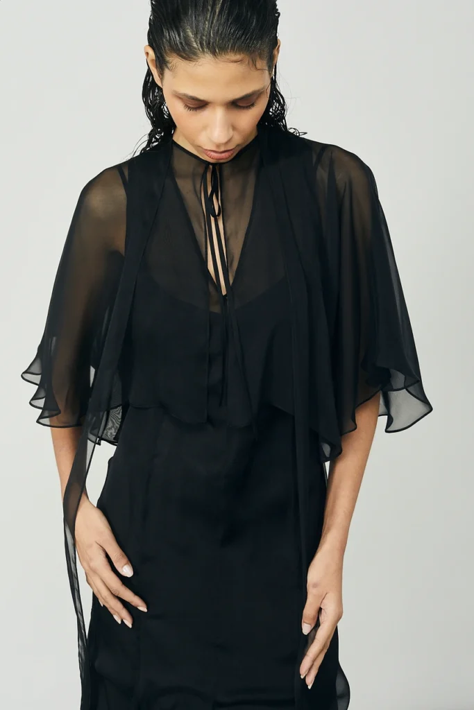Portfolio Lorenzo Berni_096 - Lorenzo Berni Woman wearing a black sheer cape over a black dress, looking downward with hands at her sides.